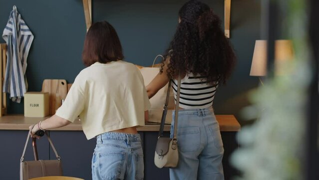 Medium back view shot of caring Biracial teenage girl helping her Caucasian female friend with cerebral palsy to unpack paper bag with food products at home after going grocery shopping together