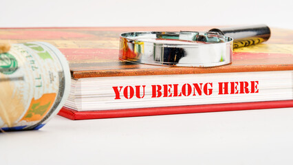 Diversity, business, inclusion and belonging concept. You belong here symbol on the pages of the book next to a roll of dollar bills and a magnifying glass on the book on a white background