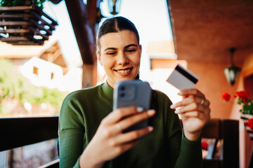 One young caucasian woman is buying or paying online on her mobile phone using credit card on her balcony 
