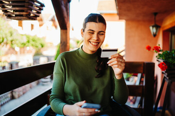 One young caucasian woman is buying or paying online on her mobile phone using credit card on her balcony 
