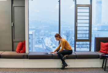 A businesswoman utilizes her laptop while seated by the window of a large corporate building,...