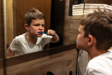 Caucasian young boy is standing in front of a mirror, diligently brushing his teeth. He is focused...