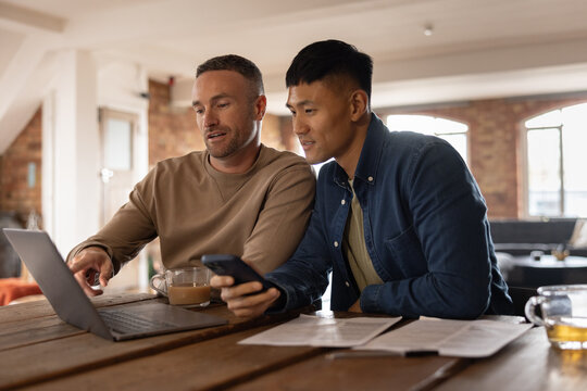 Gay couple sitting in kitchen using a laptop and smartphone for personal finances