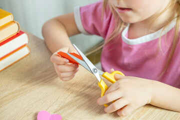 left-handed little girl learning to cut paper with scissors, making shapes like a house, heart and...