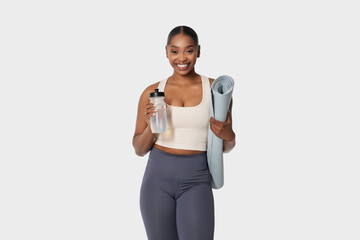 African American woman standing while holding a yoga mat in one hand and a water bottle in the...