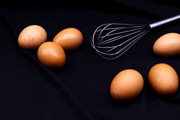 Eggs and steel whisk placed on black background. Egg beater for whisking, still-life food with a...