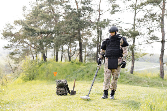 A man in a military uniform and bulletproof vest works in the forest with a metal detector. A minesweeper performs work on demining the territory