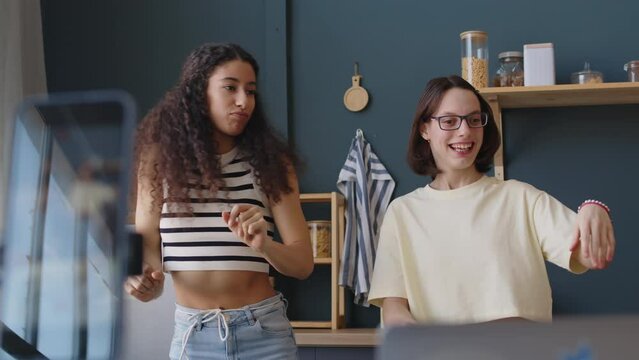Medium shot of joyful Caucasian teenage girl with cerebral palsy filming dance clip for social media with Biracial female friend on smartphone while spending leisure time at home together
