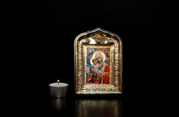 A lighted candle next to the icon of the Mother of God with the baby Jesus in her arms, on a black background. Decorative or ornamental image