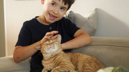 The young boy is using his hand to gently pet the orange cat on the couch