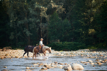 Cowgirl crossing the river riding a horse and leading a mule