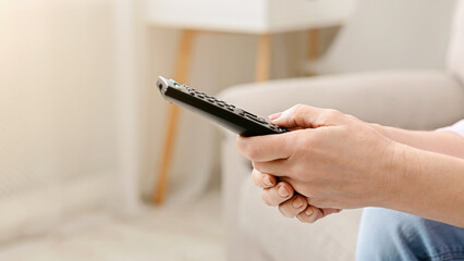 Cropped of woman is seated on a couch, holding a remote control in hand, appear to be watching television or adjusting settings on a device