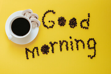 A white coffee cup and saucer stand on a yellow background. The saucer contains slices of champignon mushrooms. Nearby is a good morning phrase made from coffee beans. mushroom coffee