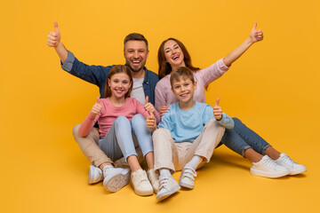 A smiling family consisting of a mother, father, daughter, and son are sitting close to each other against a vibrant yellow backdrop, all giving a thumbs-up sign in a show of happiness and unity. - Powered by Adobe