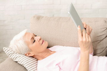 Elderly woman is seen laying on a couch, holding a tablet in her hands. She appears relaxed as she...