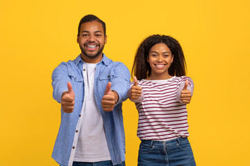 African American man and woman are smiling and showing thumbs up in approval. They are dressed casually and standing against a plain background, emphasizing their positive gesture.