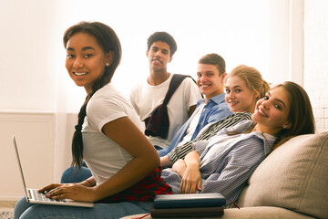 A diverse group of friends seated together on a couch inside a room. They are focused on a laptop...