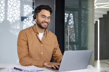Smiling customer service representative working at laptop with headset in modern office