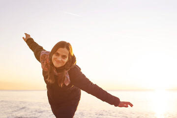 Young, playful, happy woman spread her arms in a flying gesture on the beach by the sea at sunset in spring