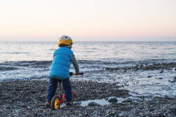 A small child in a blue jacket, in a bicycle helmet and with a red balance bike on a pebble beach by the sea at sunset, rear view