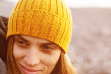 Portrait of a young woman in a yellow knitted hat and scarf on the beach near the sea in the evening at sunset in winter, close-up, soft selective focus