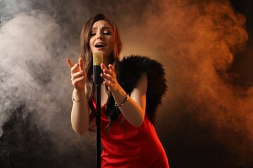 Beautiful young woman in stylish dress with microphone singing on dark background in color lights...
