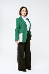 Attractive middle-aged business woman in black trousers and a green jacket stands with a laptop on a white background. Full-length portrait of a business lady.