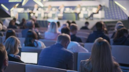 A view from the back of a conference hall showing an attentive audience watching a presentation at a modern venue with blurred foreground.