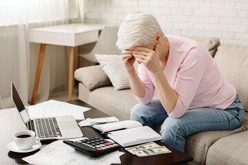 A senior woman is seated on a couch with her hands covering her face, showing signs of stress or headache, surrounded by financial documents, a calculator, money, and a laptop computer