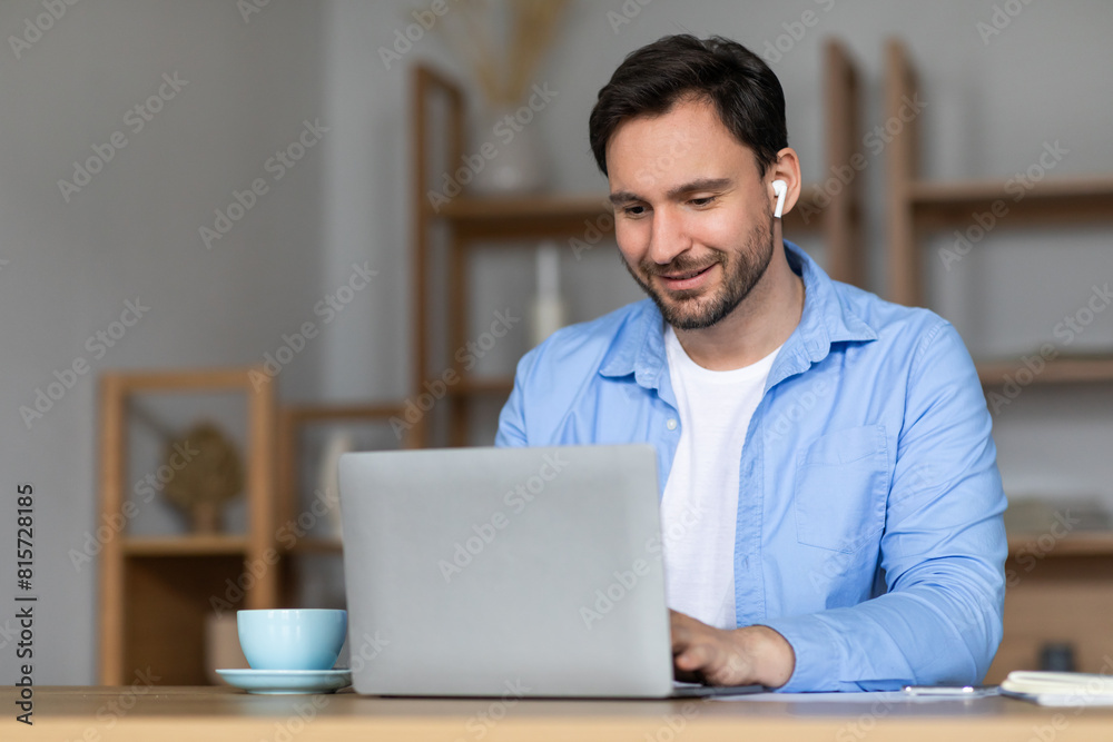 Aufkleber A man is seated at a table, working on a laptop, using earbuds. He is focused on the screen, typing and browsing. The room is simple and functional, with a few items scattered around.