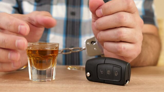 A man in handcuffs drinks a glass of alcohol and takes the car keys. The concept of driving under the influence of alcohol.