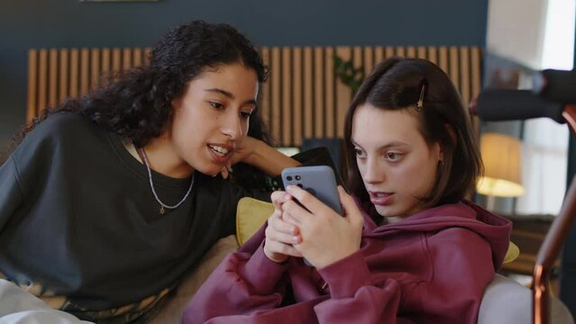 Medium shot of Caucasian teenage girl with cerebral palsy browsing on smartphone and chatting with Biracial female friend while chilling at home together after school