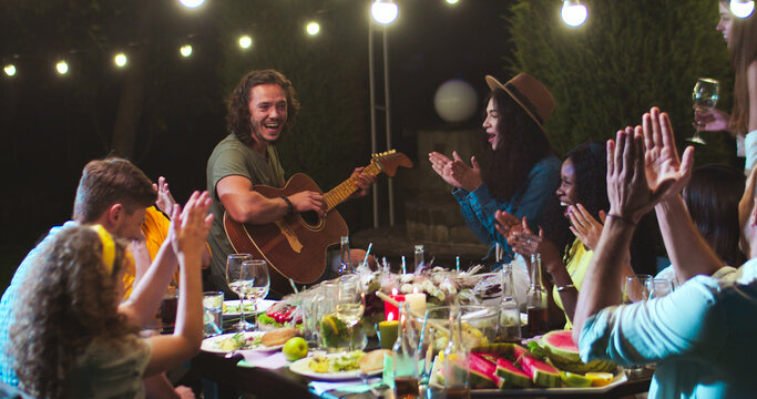 Joyful mixed-race young people having fun at party in garden at summer evening. Happy Caucasian male playing the guitar and singing outdoor. African American and Asian girls applaud. Leisure concept