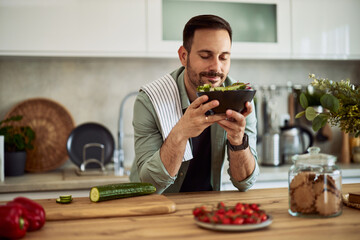 A happy adult man holding a bowl and smelling the healthy salad in the kitchen