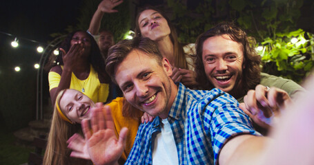 POV of joyful Caucasian man talking on video outdoor. Beautiful happy African American girl smiling. Handsome men waving their hands. Multi-ethnic friends at garden party in evening. Fun concept