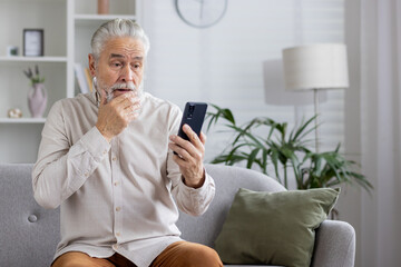 Concerned senior man looking at smartphone while sitting on sofa in living room