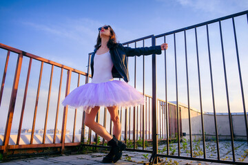 a ballerina in tutu stands at the fence on the roof .at sunset in boots and a jacket