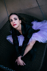 a ballerina in a tutu and a black jacket with red lipstick is sitting on a swing with a pained look
