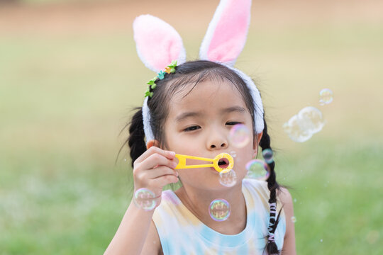 Happy 6 year old Asian little girl with bunny ears blowing soap bubbles in park, having fun, sunny day. Portrait of cute child playing outdoors in summer in nature