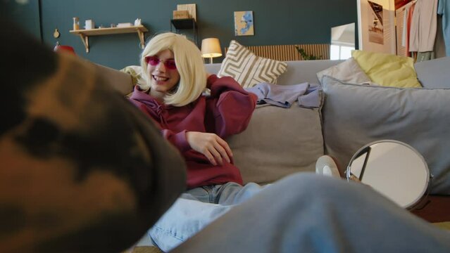 Stab shot of Caucasian teenage girl with cerebral palsy wearing blond wig and pink y2k glasses sitting on floor in modern living room dancing and chatting with friend, chilling together indoors