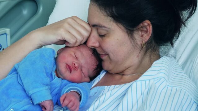 New 30s mother kissing newborn baby on forehead bonding with infant right after childbirth resting at hospital bed during the initial days of son's life, motherhood and maternal care concept