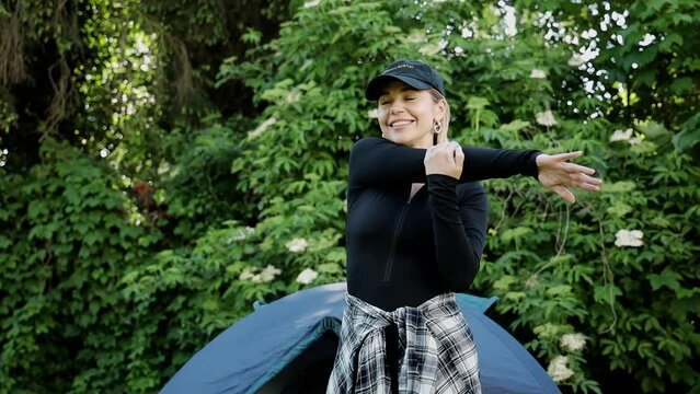 a girl doing morning exercises in nature. She stands in front of a tent surrounded by greenery and permanent trees. The morning sun shines softly, creating a warm and calm atmosphere. The girl is dres