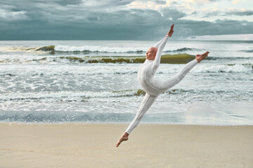 Young hairless ballerina with alopecia in white futuristic suit dancing and jumping on sea sandy beach, metaphoric surreal scene with bald pretty teenage girl exudes confidence and unique beauty