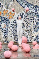 Outdoor dance of hairless girl ballerina with alopecia in white futuristic suit, standing and holding two pink spheres on abstract mosaic Soviet background, symbolizes self expression