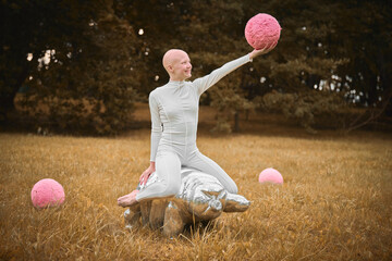 Young hairless girl with alopecia in white cloth sits on tardigrade figure and holds pink ball in hand on fall lawn park, surreal scene with bald teenage girl engages with symbolic elements