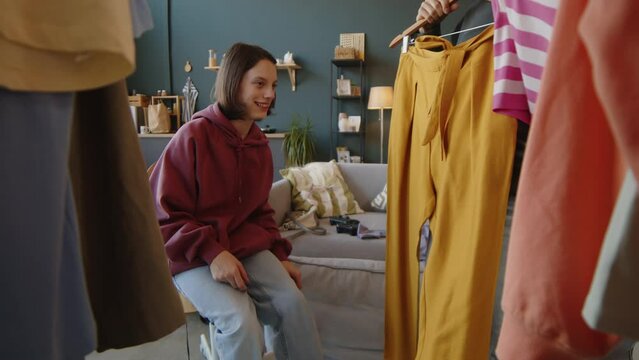 Medium shot of Caucasian teen girl with cerebral palsy sitting on chair by clothes rack in modern bedroom and helping Biracial female friend to choose outfit to go out