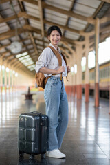 Happy young Asian female tourist carrying a backpack, holding a camera, preparing to wait for the train at the train station waiting for her vacation trip.