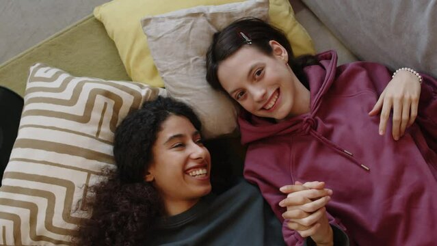 High angle arc portrait shot of Caucasian girl with cerebral palsy and her Biracial female friend looking at camera and smiling while lying on couch holding hands