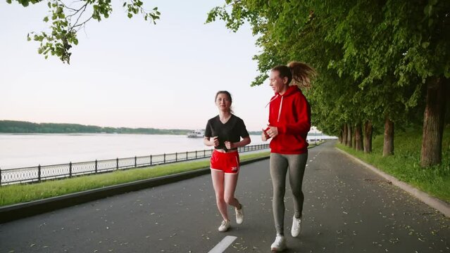 wo pretty women communicate with each other while doing sports. Two young girls in red clothes running in the park. Spend time with friends. TMorning or evening jogging along the river embankment.