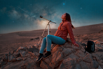 beautiful woman sitting in the desert alone next to a telescope at night watching the starry sky,...
